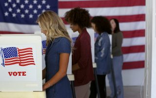 Four women of various demographics filling in ballots and casting votes in booths at polling station, US flag on wall at back.