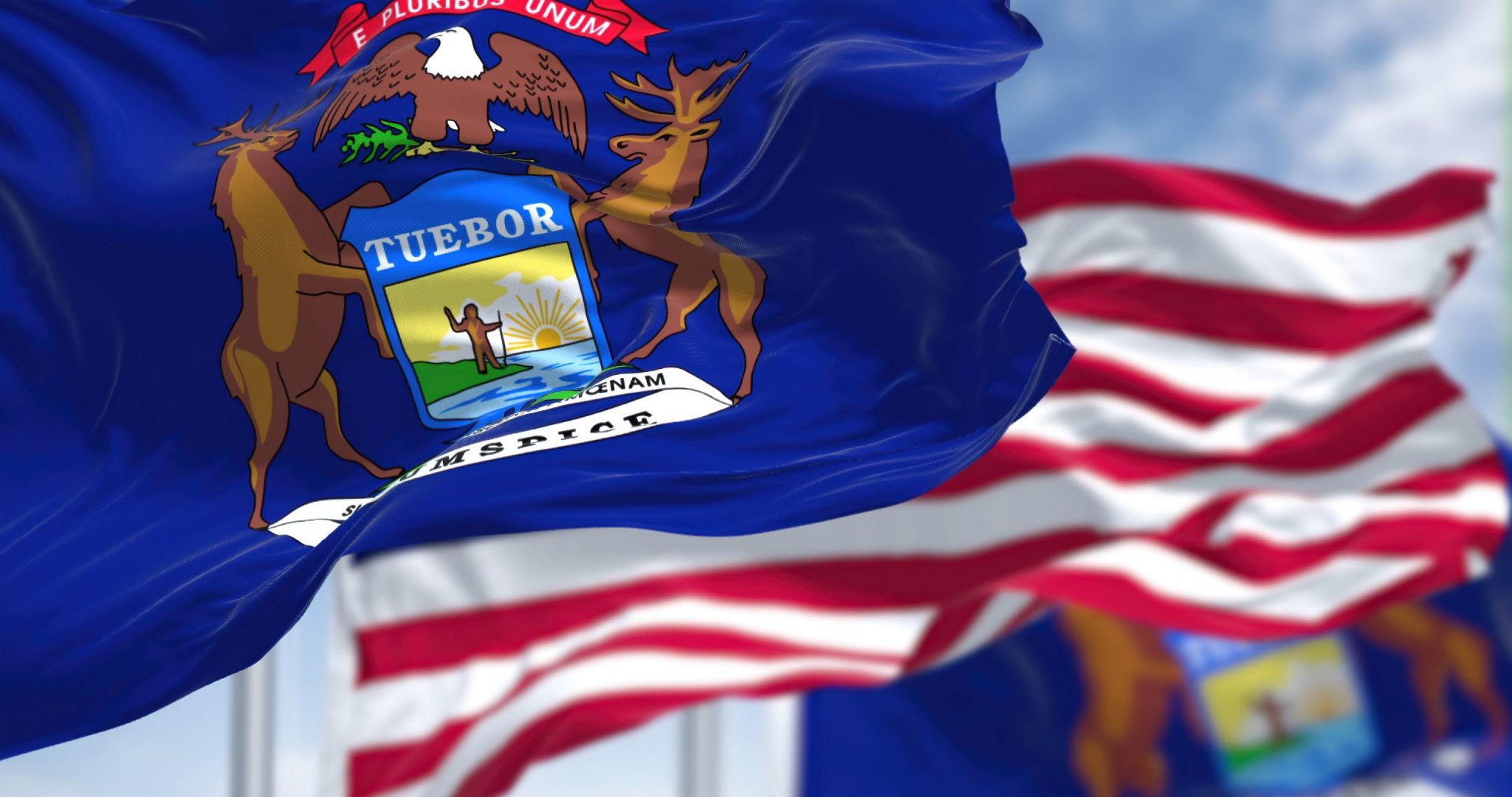 The Michigan state flag waving along with the national flag of the United States of America. In the background there is a clear sky. Michigan is a state in the upper Midwestern of United States